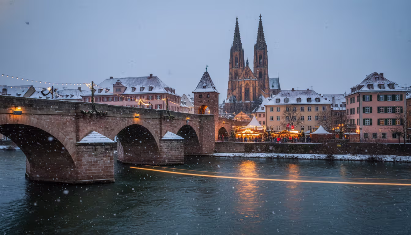 Medieval stone bridge in Regensburg in winter with Gothic cathedral and Christmas market stalls illuminated at dusk