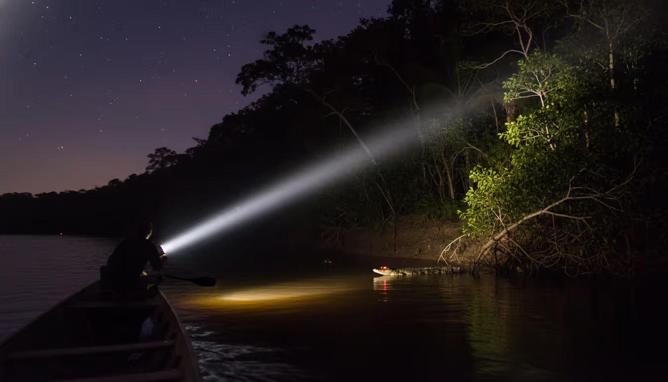 Nighttime Amazon expedition with flashlight beam illuminating caiman eyes glowing red on a dark riverbank