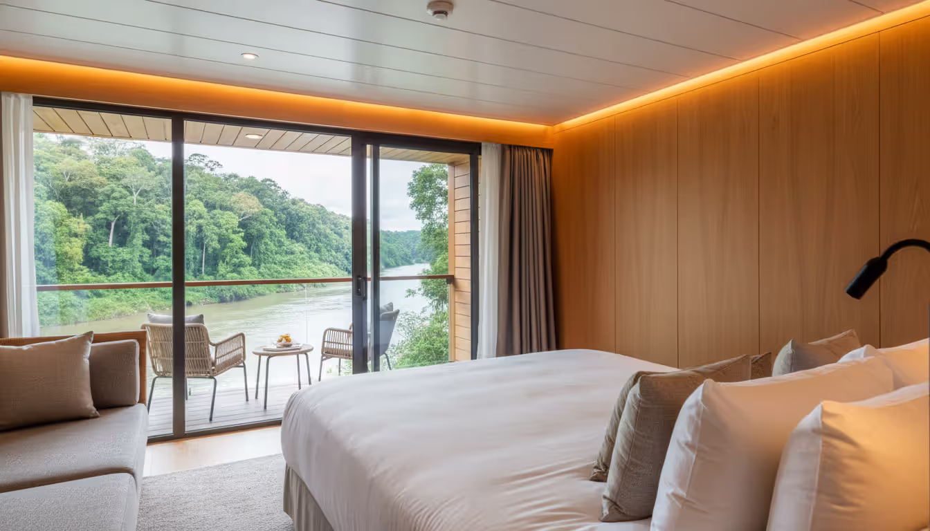 Luxury Amazon cruise ship suite interior with floor-to-ceiling panoramic windows showing tropical rainforest and river view with private balcony