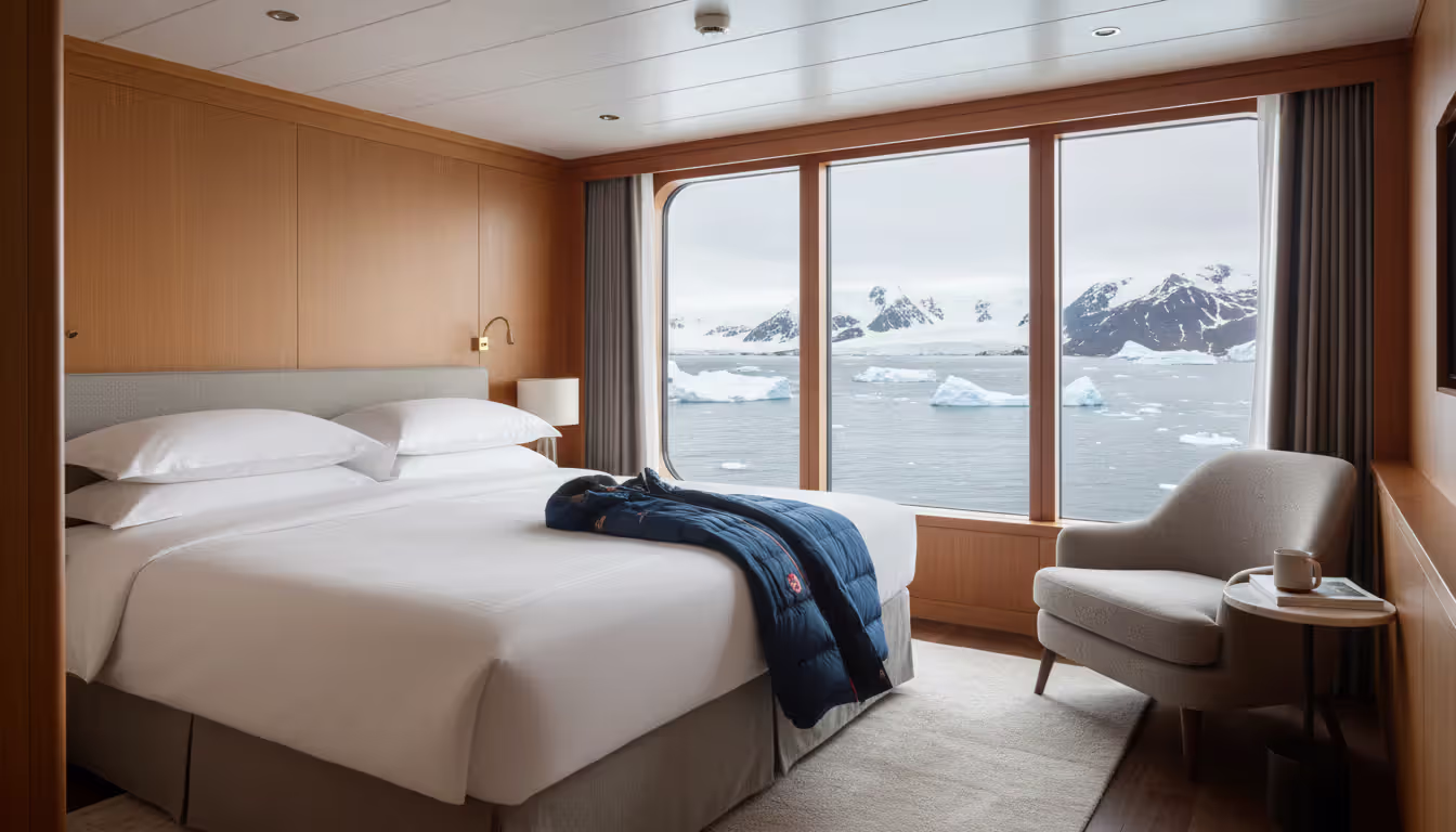 Spacious luxury suite cabin on an expedition ship with floor-to-ceiling window showing Antarctic ice and mountain scenery outside