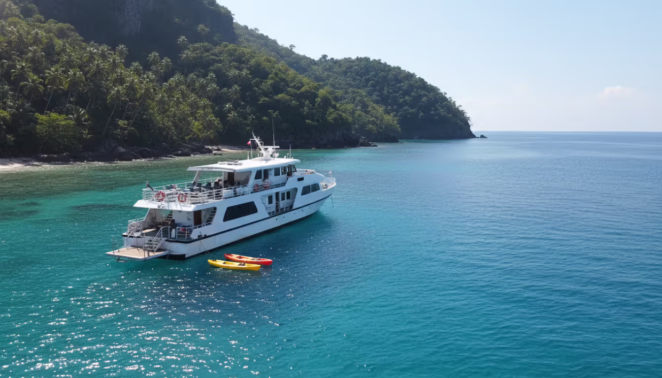 Small white luxury cruise ship anchored in a secluded turquoise bay with a watersports platform and colorful kayaks near a tropical island