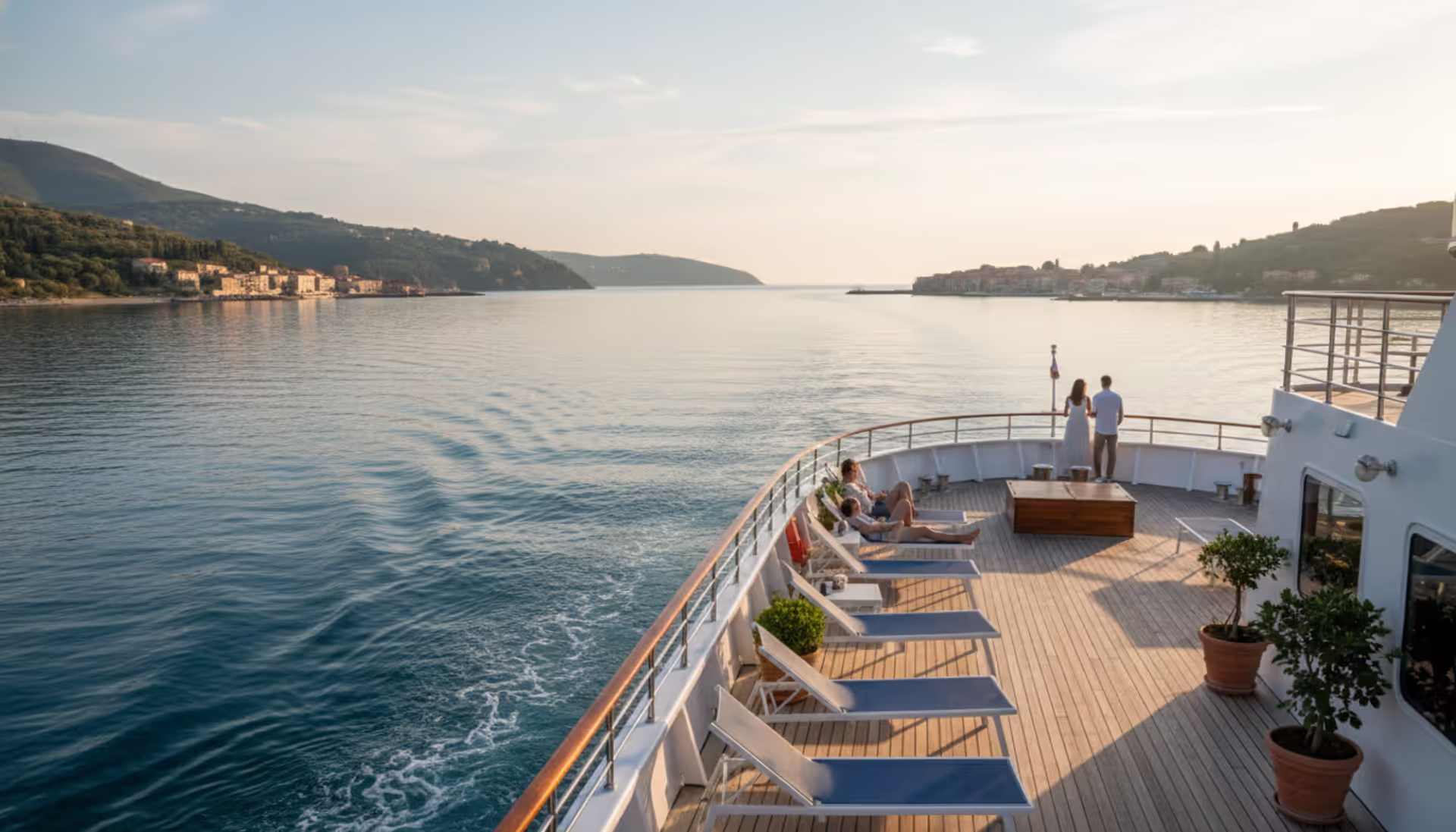 Elegant small luxury cruise ship sailing along a scenic Mediterranean coastline with a coastal town and green hills in the background on a sunny morning