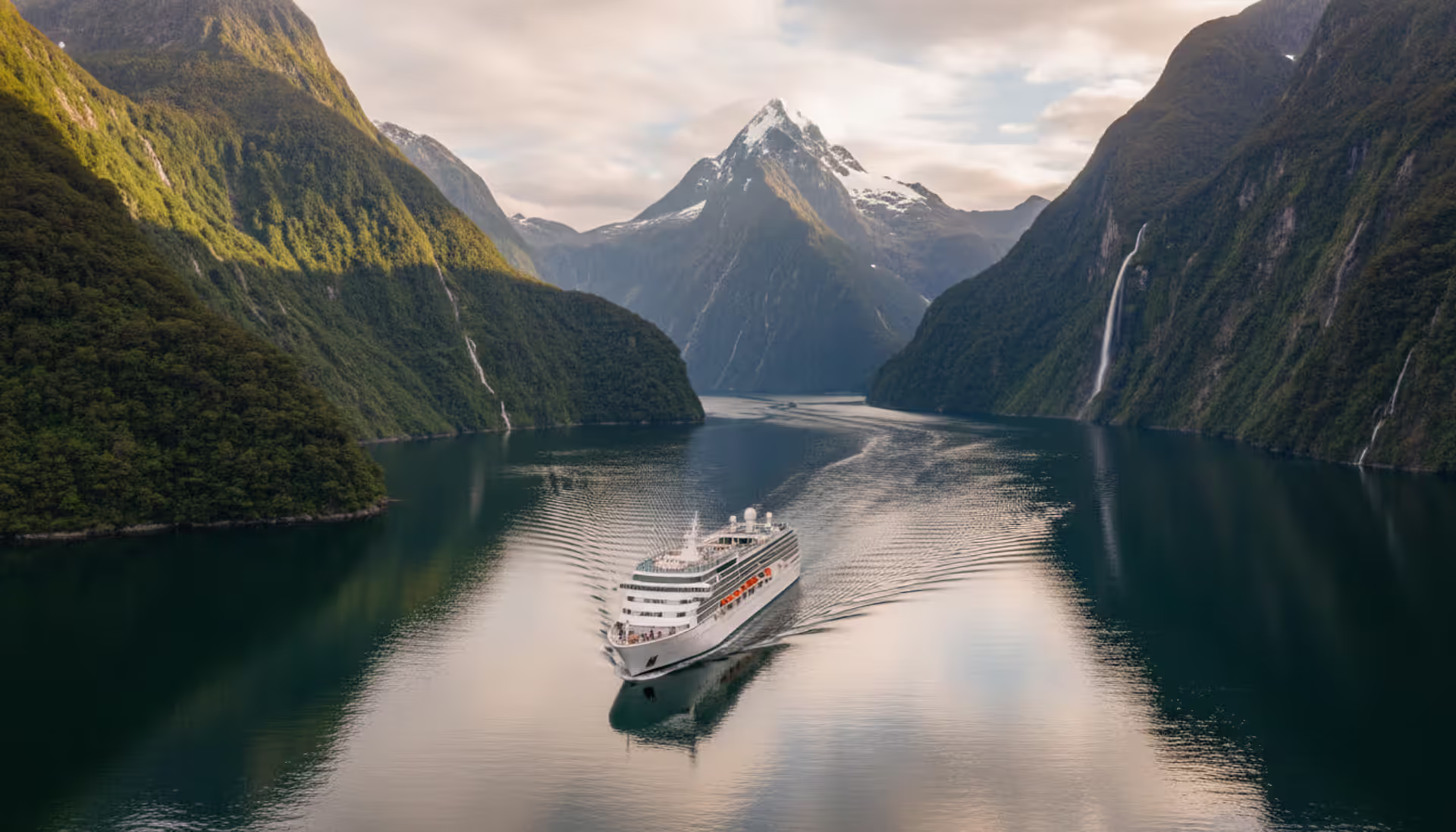 Luxury white cruise ship sailing through Milford Sound fjord in New Zealand with towering green cliffs and Mitre Peak in the background