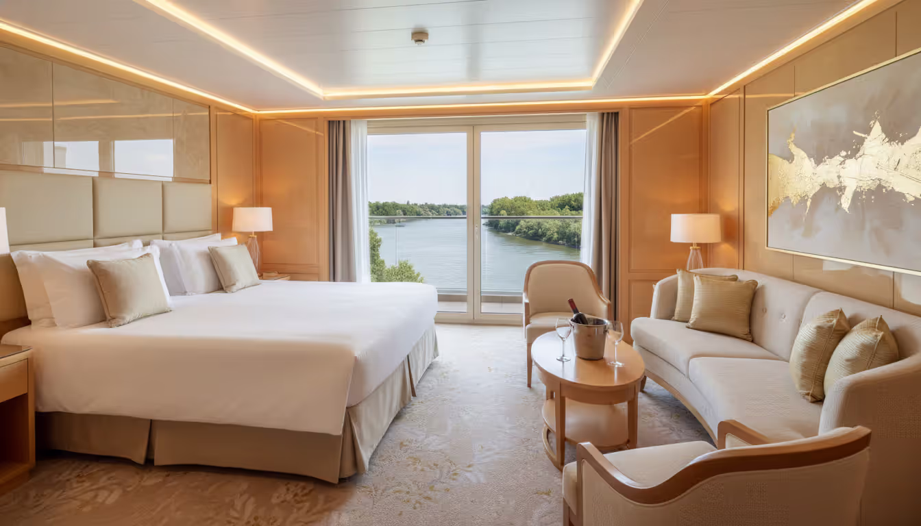 Luxurious river cruise suite interior with floor-to-ceiling glass doors overlooking a river, king-size bed, and elegant sitting area