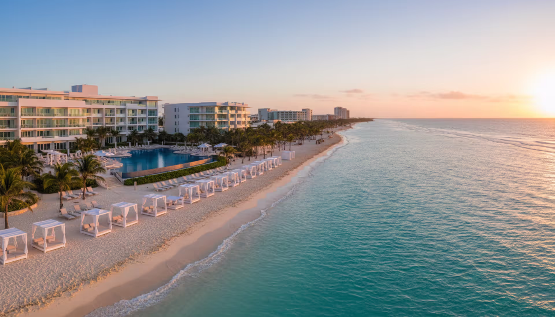 Panoramic view of a luxury all-inclusive resort on the Mexican Caribbean coast with turquoise sea, white sand beach, elegant sunbeds, infinity pool, and tropical vegetation at sunset