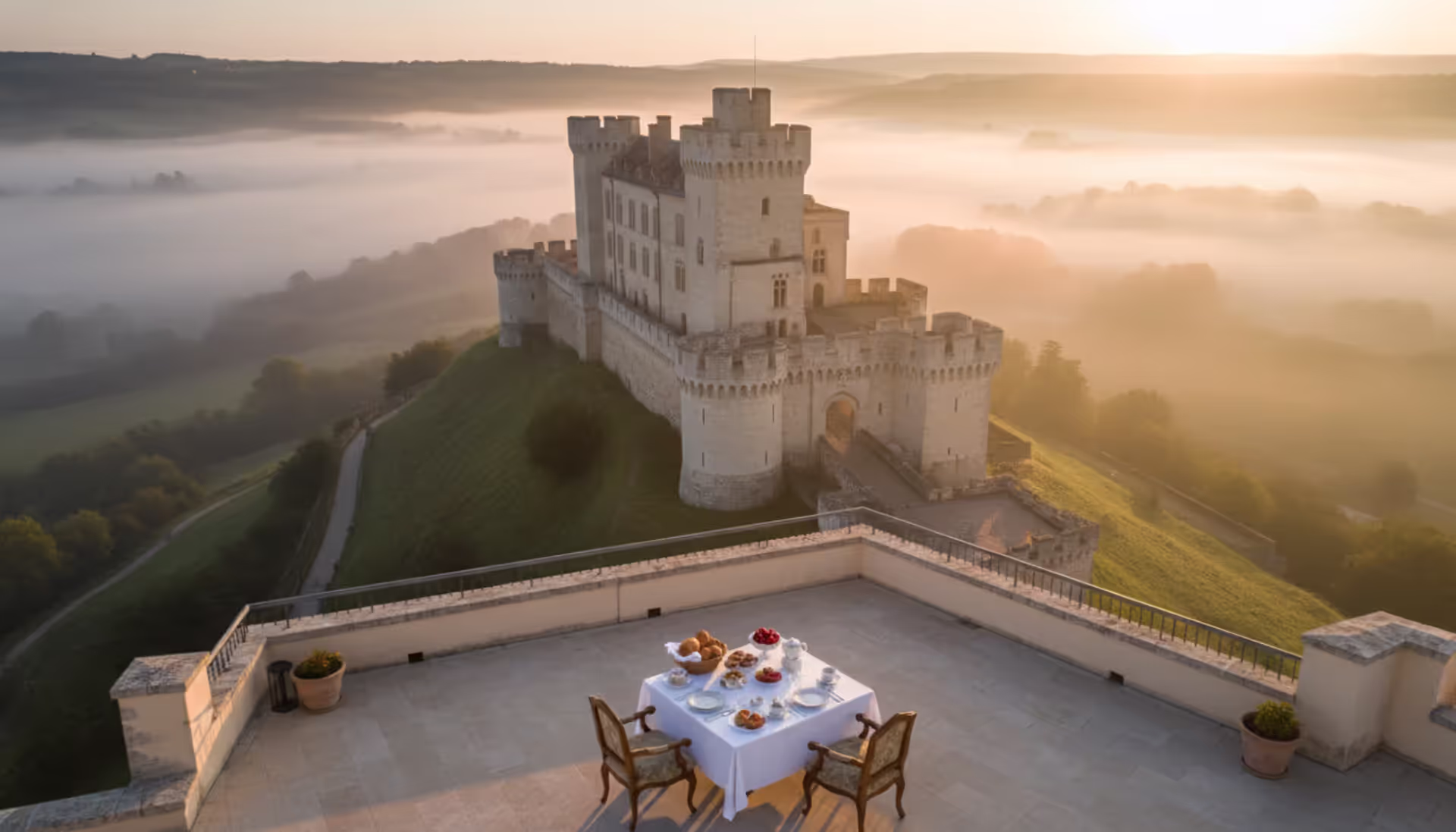 Luxury breakfast terrace overlooking a medieval European hilltop castle surrounded by morning mist and green valley