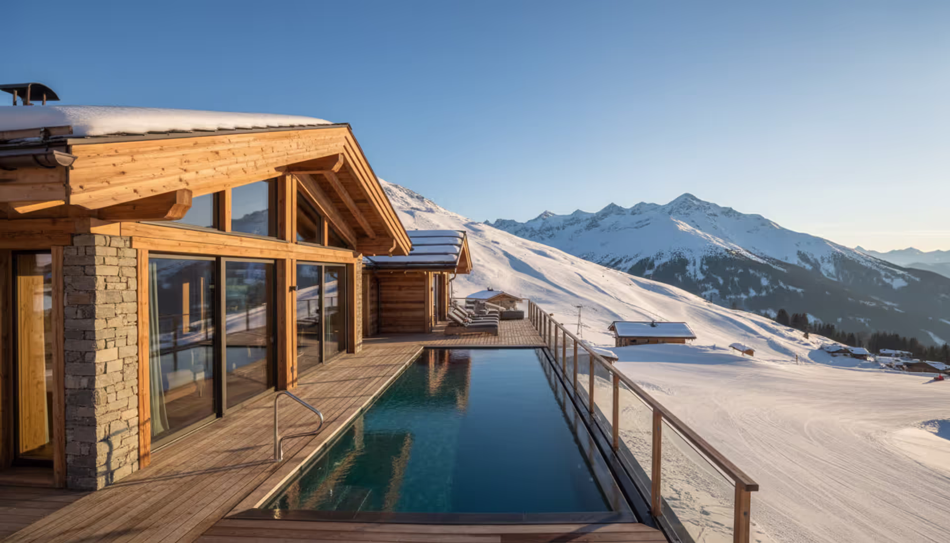 Panoramic sunrise view of a luxury alpine ski resort with a wooden chalet, heated outdoor pool, and groomed ski slopes surrounded by snowy mountain peaks