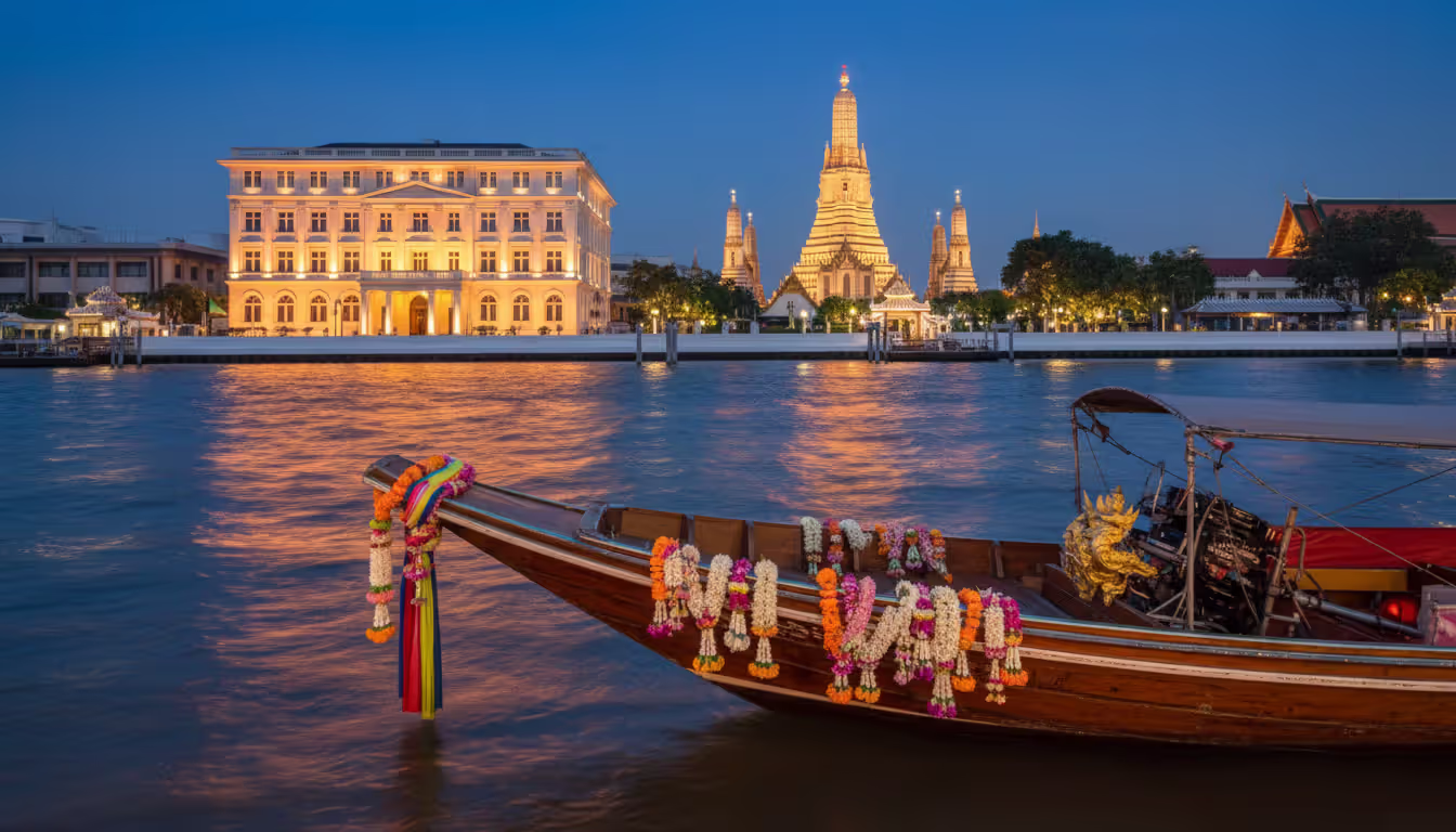 Evening view of Bangkok Chao Phraya River waterfront with an ornate longtail boat, illuminated colonial heritage hotel, and golden Buddhist temple spires reflecting in the water