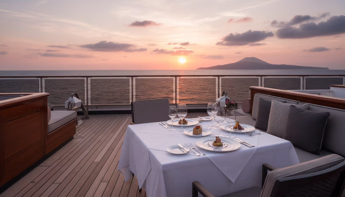 Elegant outdoor dinner setup on a luxury cruise ship deck with white tablecloth wine glasses and gourmet dishes against a dramatic pink and orange ocean sunset with a volcanic island silhouette