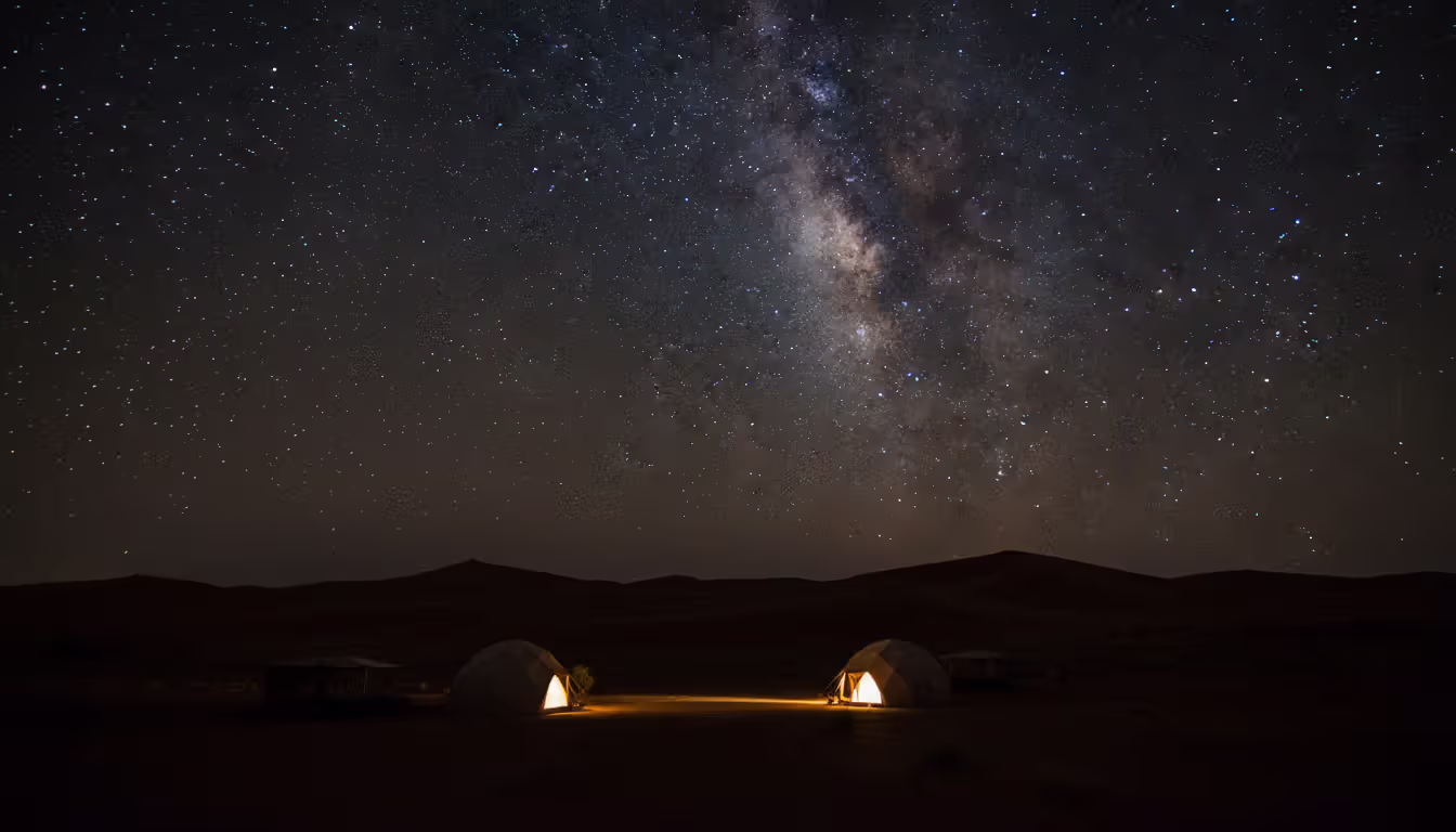 Milky Way and star-filled night sky above silhouetted Namibian desert dunes with a softly lit safari camp in the foreground