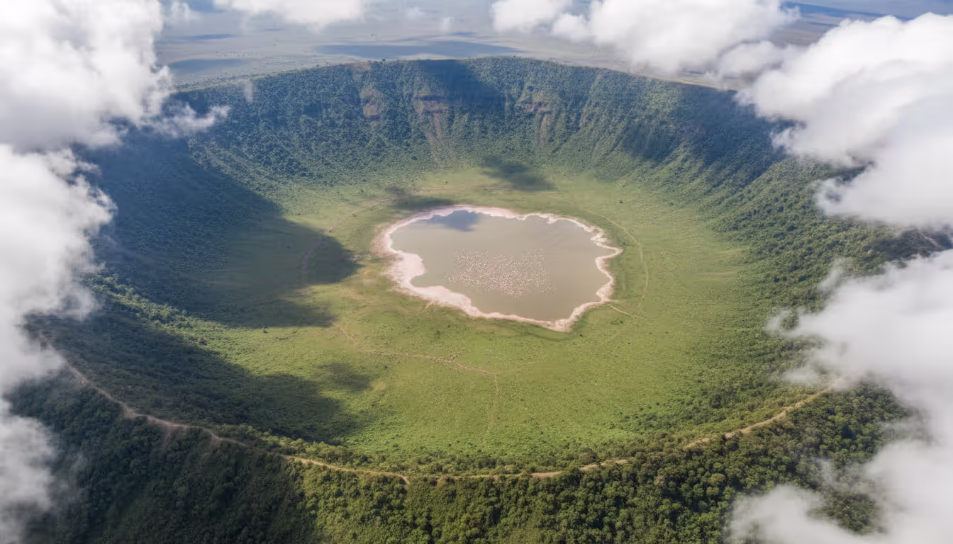 Aerial view of Ngorongoro Crater caldera with green floor, flamingo-covered lake, steep vegetated walls and clouds partially filling the basin