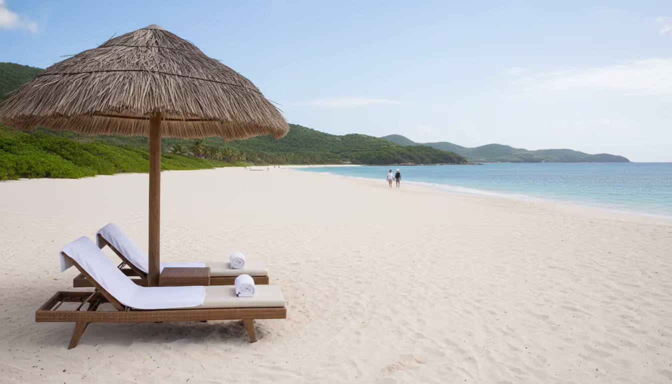A pristine white sand Caribbean beach with two sun loungers under a straw umbrella and a couple walking along the shoreline in the distance