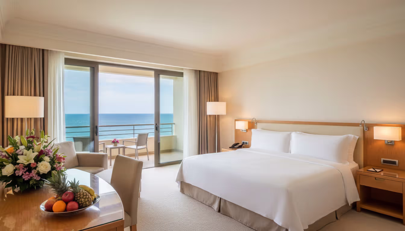Luxurious five-star hotel suite with panoramic ocean view through floor-to-ceiling glass doors with white bedding and fresh flowers