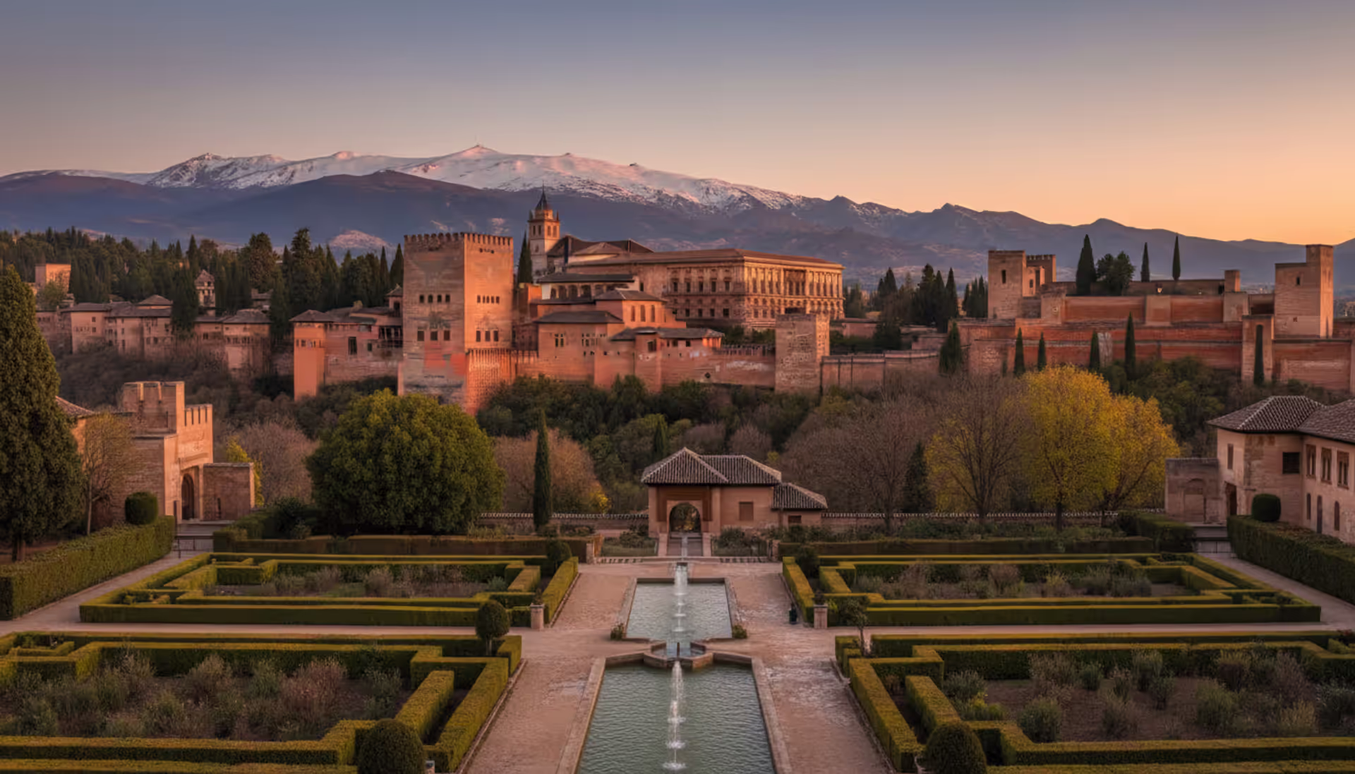 Alhambra palace in Granada Spain at sunset with manicured gardens fountain and Sierra Nevada mountains in the background