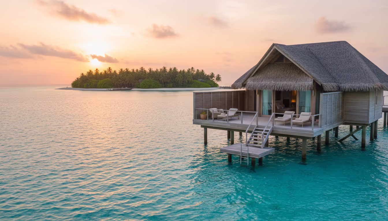 Overwater luxury bungalow on stilts above crystal-clear turquoise lagoon with wooden deck, sun loungers, steps into water, and tropical islands in the background at sunset