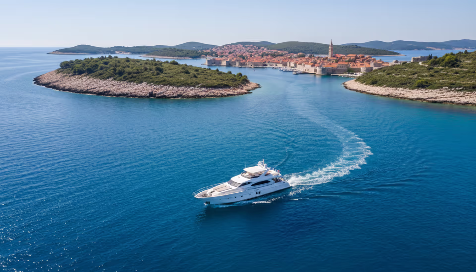 Aerial view of a luxury white yacht sailing through turquoise Adriatic waters near Croatian islands with a medieval coastal town in the background
