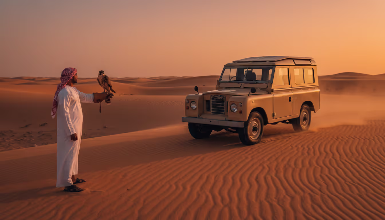 Vintage 1960s Land Rover driving through desert dunes at sunset with a falconer holding a falcon nearby