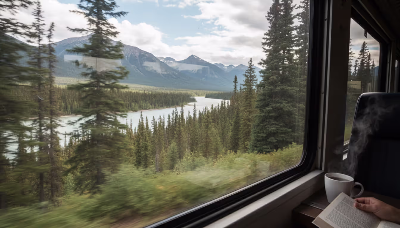 View from inside a panoramic train window looking out at vast Canadian wilderness with dense conifer forests, a wide river, and distant mountain ranges with no visible human structures