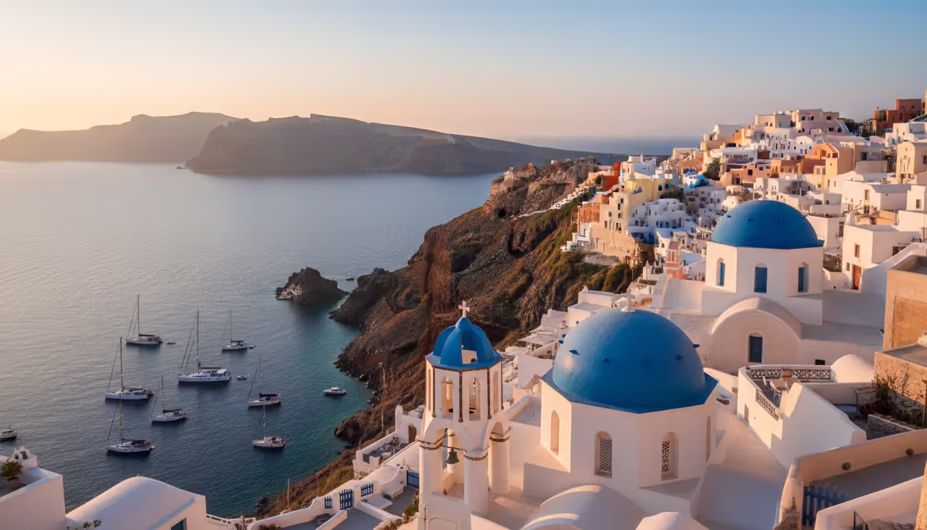 White buildings and blue domed churches of Santorini perched on cliff edge overlooking the caldera with yachts anchored below at golden hour