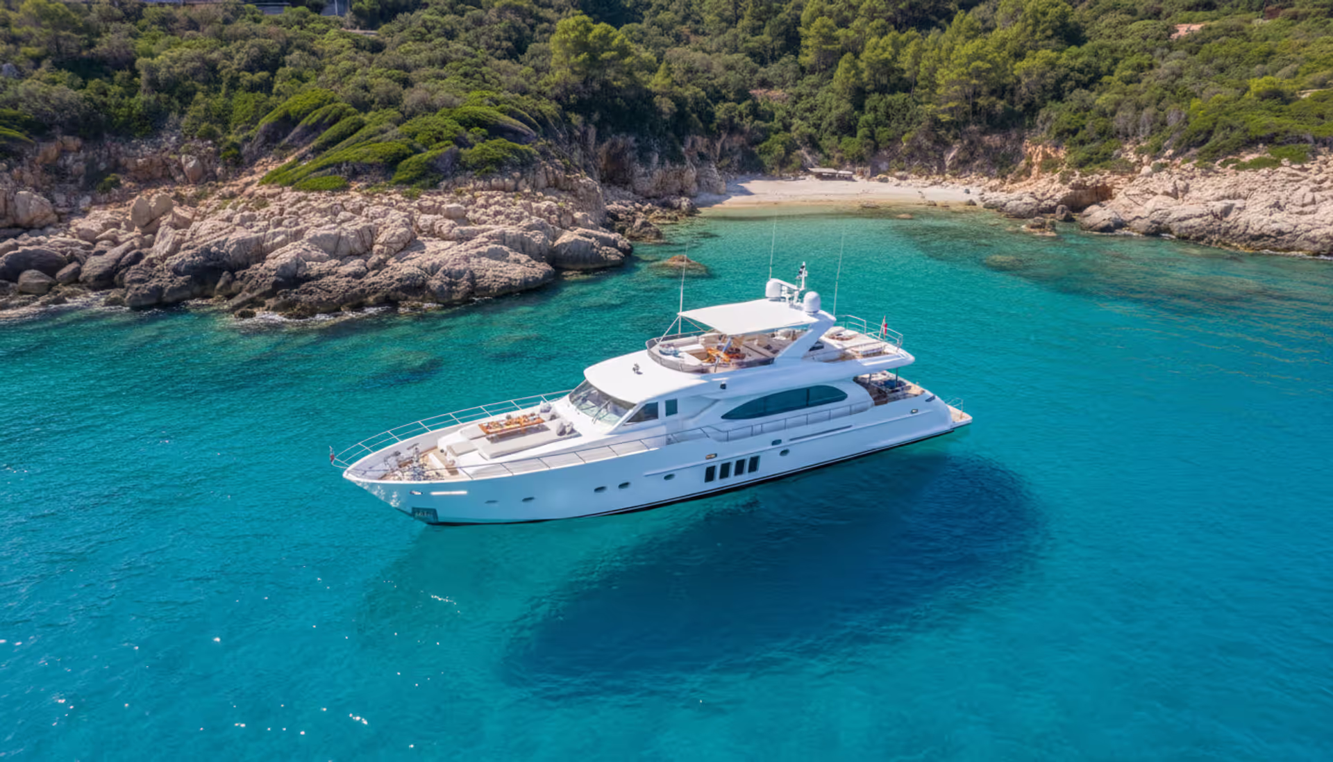 Aerial view of a luxury white motor yacht cruising along a rocky Mediterranean coastline with turquoise water on a sunny day