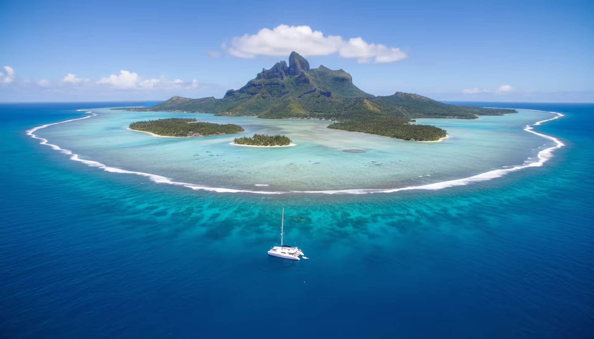 Aerial view of Bora Bora turquoise lagoon with Mount Otemanu, coral reef, and a white sailing catamaran anchored near a tropical motu
