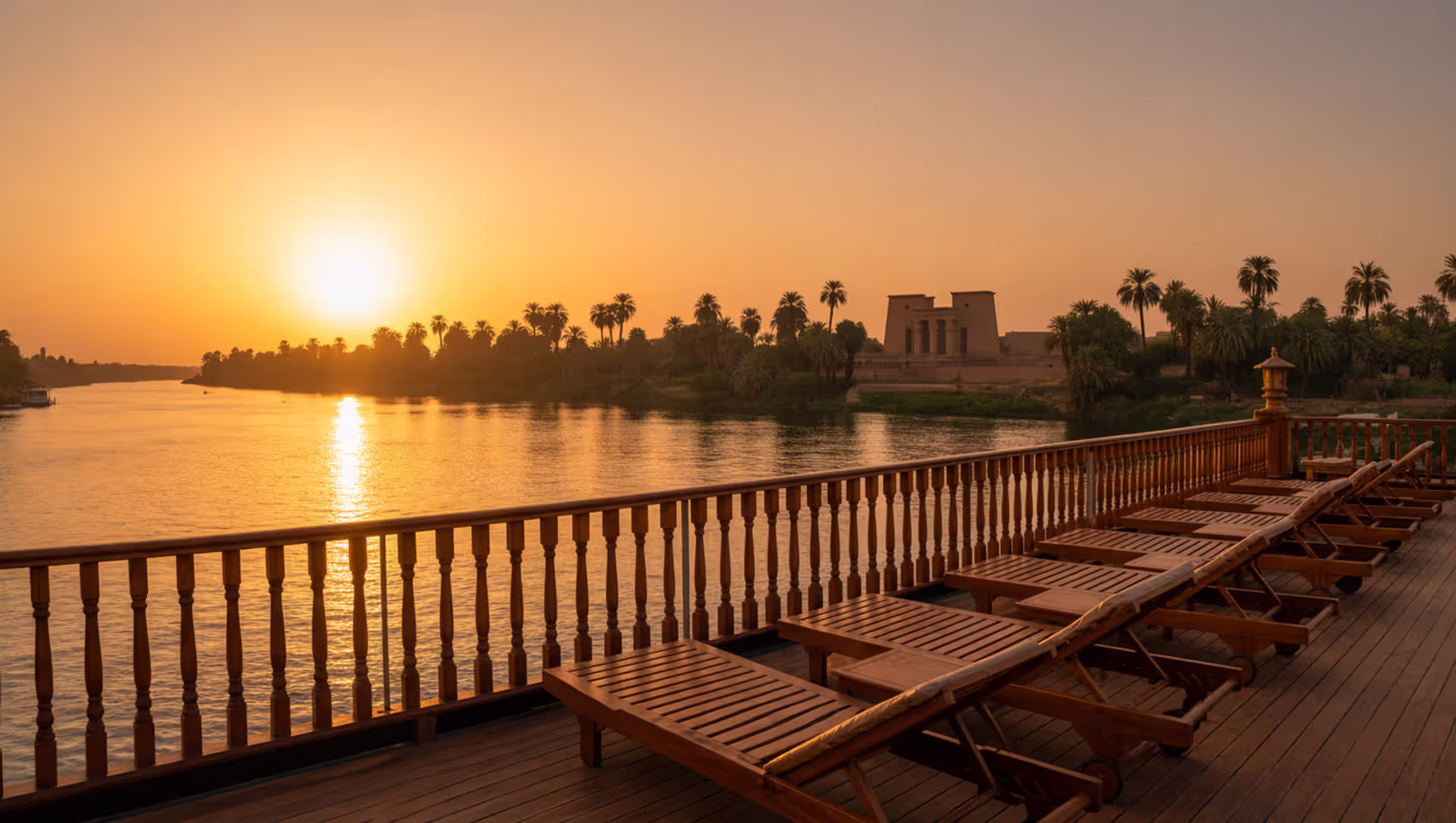 Panoramic sunset view from the deck of a Nile cruise ship with palm-fringed riverbanks and ancient temple silhouette in the distance